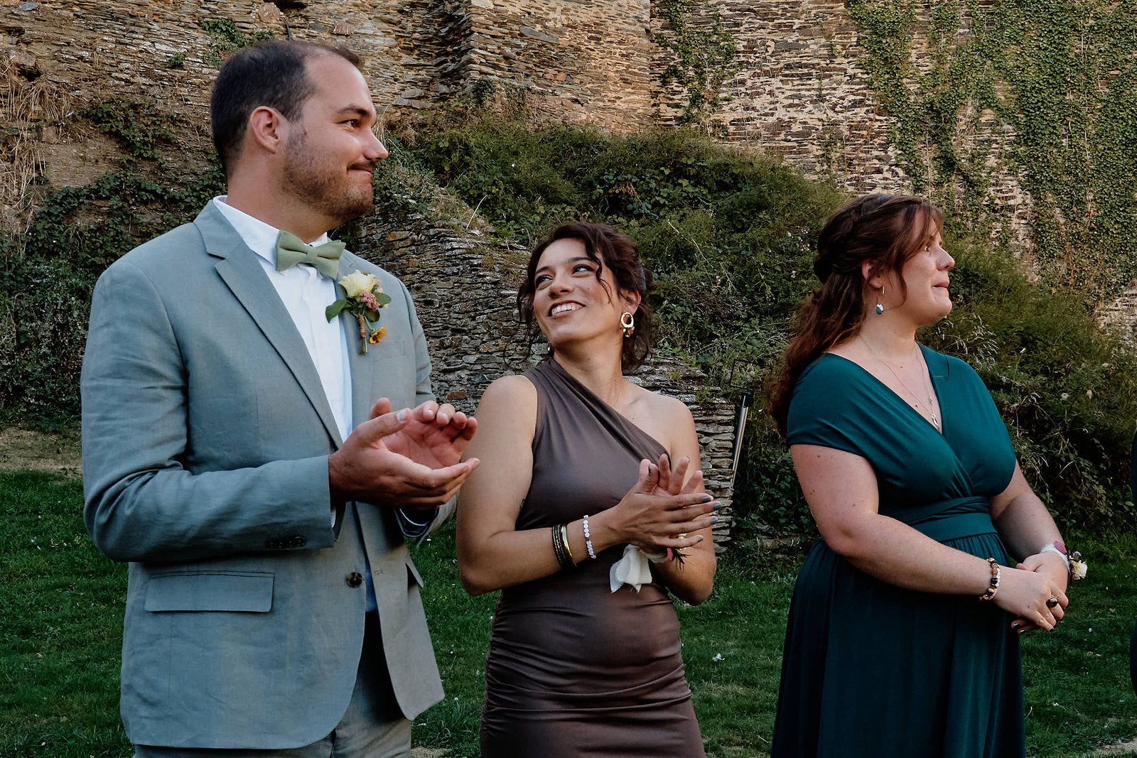 Un témoin au nœud papillon applaudit en regardant arriver la mariée, une invitée en robe marron lève les yeux vers lui en souriant