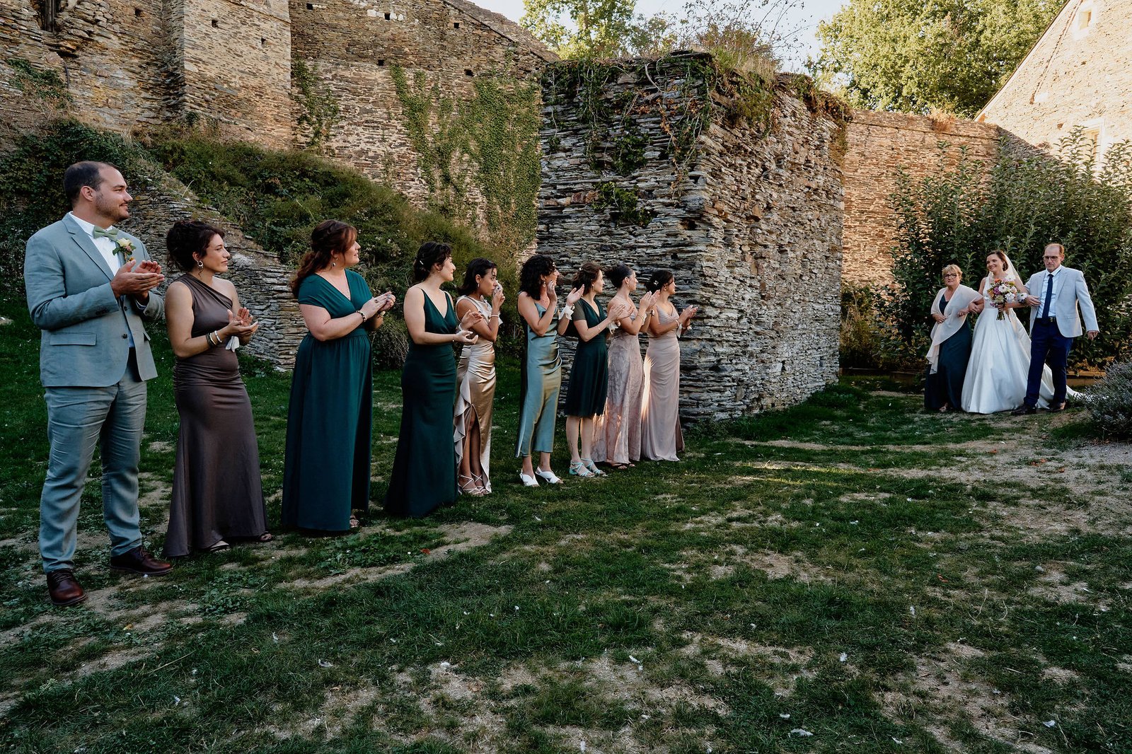 Vue d’ensemble de la cérémonie devant les remparts du Château du Plessis-Macé : le témoin et les témoins applaudissent en ligne pendant que la mariée arrive au loin avec ses parents