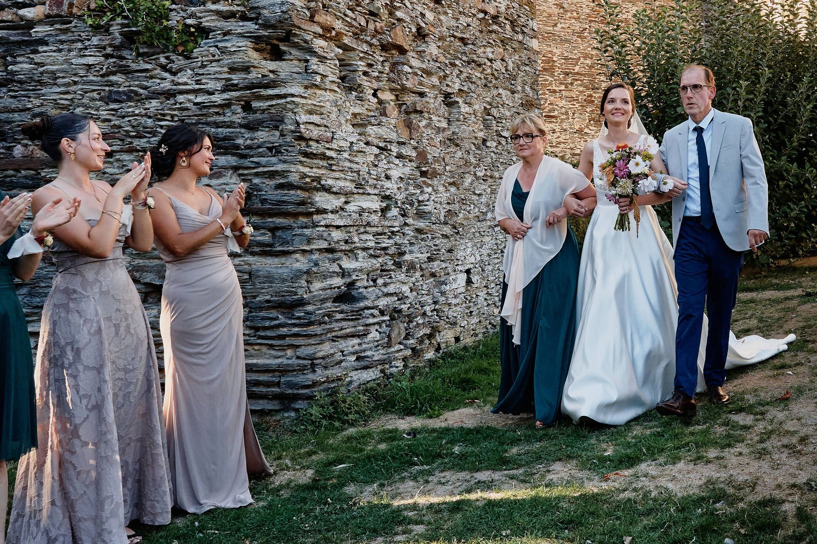 La mariée avance au bras de son père et de sa mère le long d’un mur de schiste, deux amies l’applaudissent