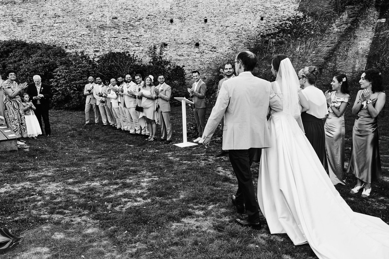 En noir et blanc, la mariée de dos au bras de son père, face à tous les invités qui applaudissent
