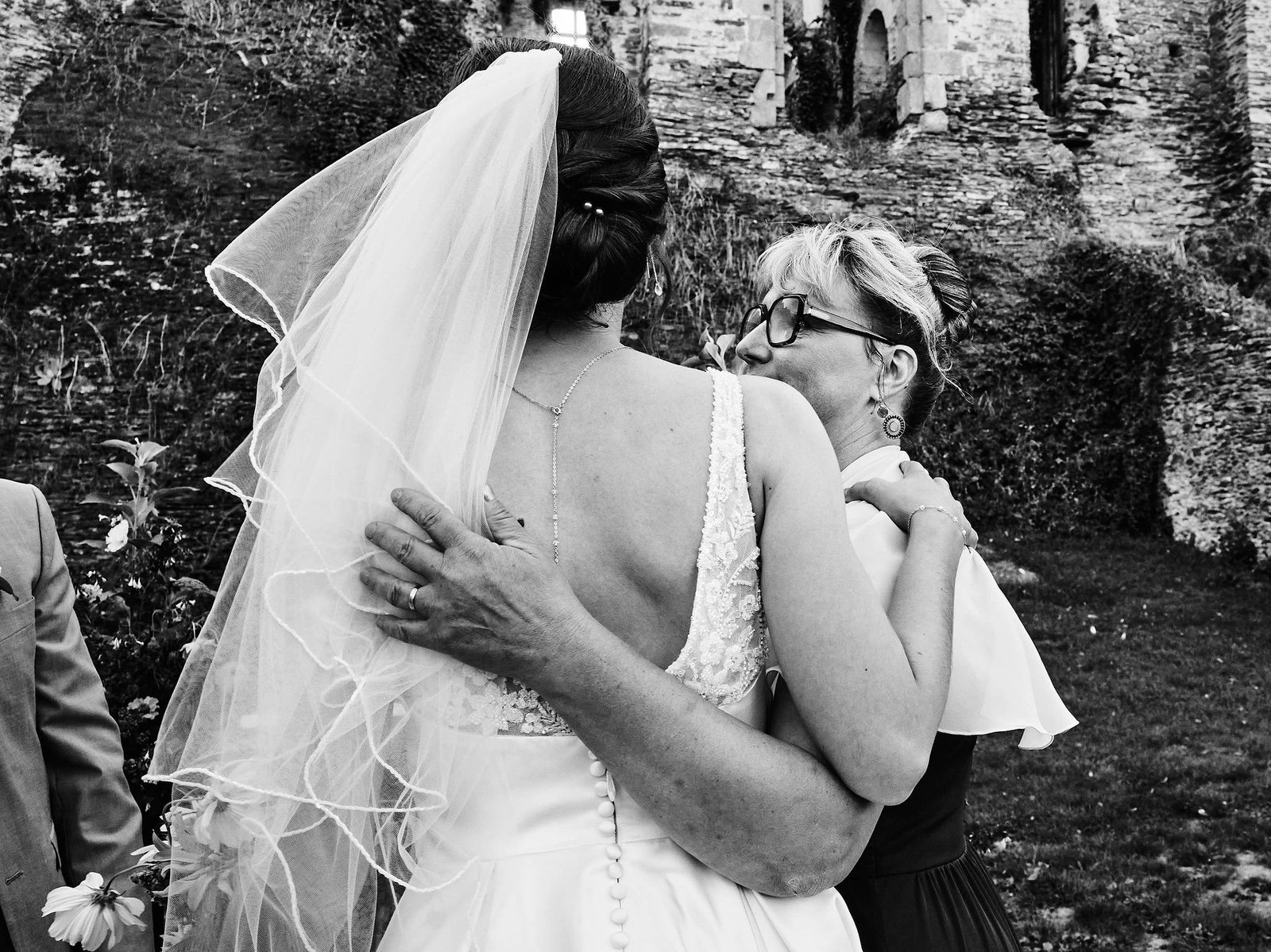 En noir et blanc, la main de la mère, alliance au doigt, posée sur le dos de la robe de la mariée pendant leur étreinte