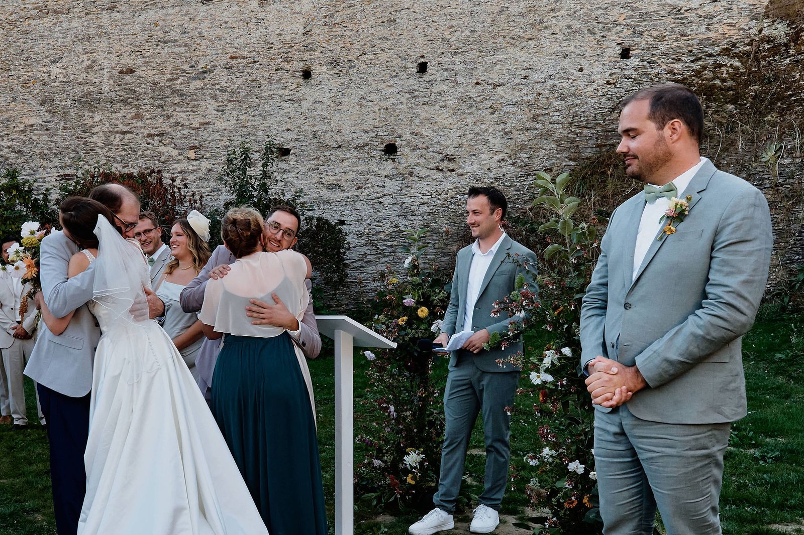 La mariée serre son père dans ses bras pendant que le marié embrasse sa belle-mère, un témoin attend à l’écart en souriant