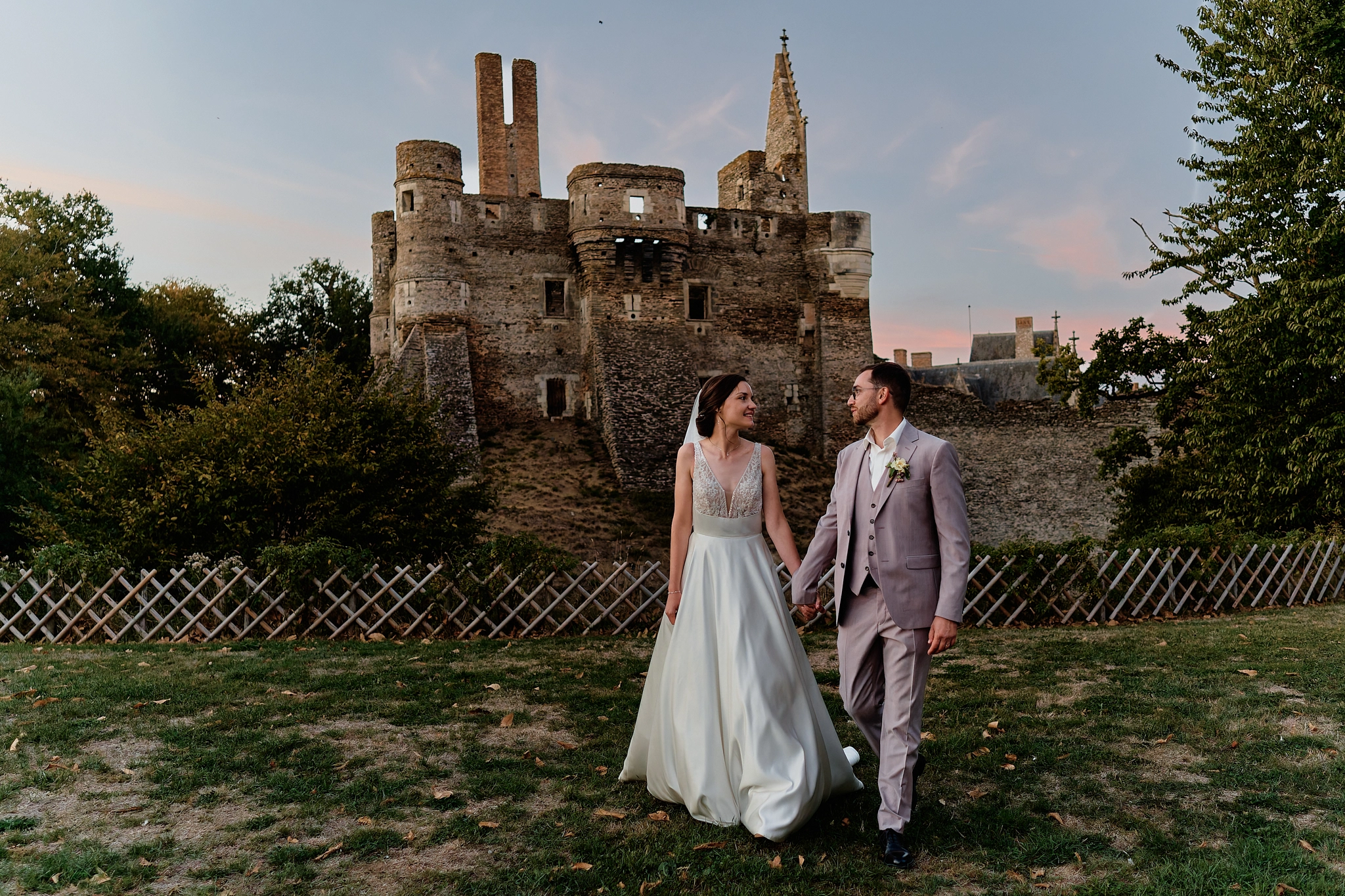 Les mariés marchent main dans la main en se regardant, devant les tours du Château du Plessis-Macé au coucher du soleil, en Anjou